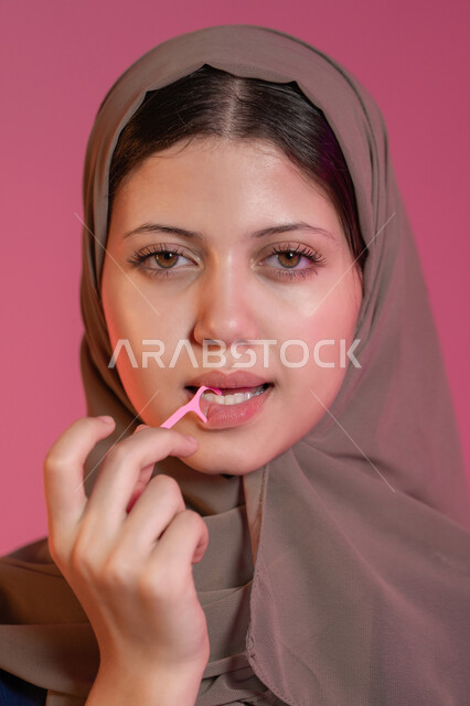 المحافظة على ابتسامة بيضاء، تجنب التسوس وأمراض اللثة، التأكد من نظافة الأسنان بعد تناول الطعام, بورتريه مقرب لامرأة عربية خليجية اماراتية محجبة تقوم بالتنظيف باستعمال الخيط السني, خلفية وردية