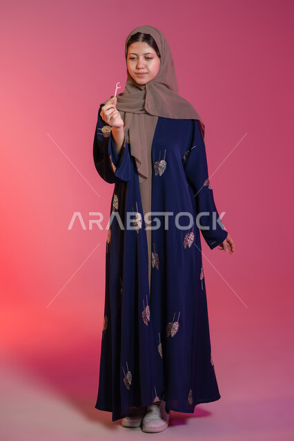 Using a toothpick, taking care of oral hygiene and health, avoiding cavities and gum disease, ensuring cleanliness after eating, portrait of a veiled Emirati Gulf Arab woman wearing a blue abaya holding a plastic toothpick in her hand, pink background