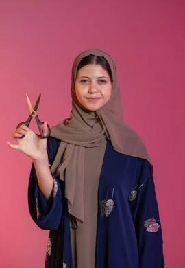 مفهوم العناية و الاهتمام بالجمال والمظهر الخارجي، قص و تصفيف الشعر في صالون الحلاقة، بورتريه لامرأة عربية خليجية اماراتية محجبة ترتدي عباءة زرقاء تحمل بيدها المقص و تنظر إلى الكاميرا بإيماءات السرور والسعادة، خلفية وردية