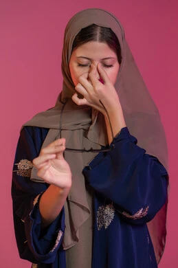 Expressions of extreme fatigue and headache, use of eyeglasses, concept of poor vision, portrait of a veiled Emirati Gulf Arab woman wearing a blue abaya, putting her hands on her face and feeling dizzy, vision correction and improvement, pink background