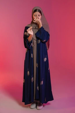 Migraine pain, feeling of headache and dizziness, need for treatment and rest, use of eyeglasses, concept of poor vision, portrait of a veiled Emirati Gulf Arab woman wearing a blue abaya, putting her hands on her face and feeling dizzy, correcting and improving vision, pink background