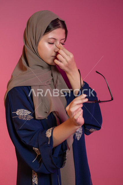 الإحساس بوجع الرأس و الدوخة, الحاجة للعلاج والراحة, الرؤية من خلال النظارة الطبية، مفهوم ضعف البصر, بورتريه لامرأة عربية خليجية اماراتية محجبة ترتدي عباءة زرقاء تضع يديها على وجهها و تشعر بالصداع, تصحيح وتحسين البصر، خلفية وردية