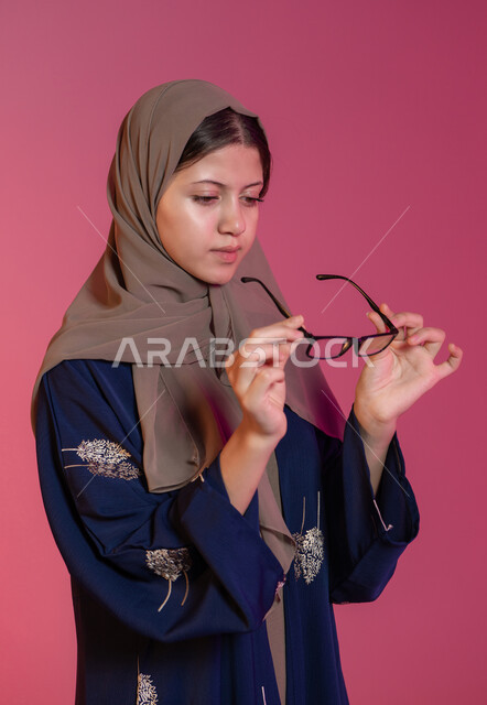 اختيار نظارات طبية مناسبة، مفهوم ضعف البصر، بورتريه لامرأة عربية خليجية اماراتية محجبة ترتدي عباءة زرقاء تمسك نظارة ملائمة لملامح وجها، تصحيح وتحسين الرؤية، متعة التسوق في الإمارات، خلفية وردية