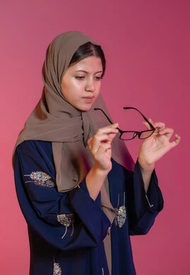 Choosing suitable eyeglasses, concept of visual impairment, portrait of a veiled Emirati Gulf Arab woman wearing a blue abaya holding glasses that fit her facial features, correcting and improving vision, the pleasure of shopping in the Emirates, pink background