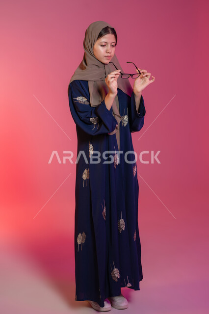 Wearing glasses for poor eyesight, taking care of one's appearance, the concept of poor eyesight, a portrait of a veiled Emirati Gulf Arab woman wearing a blue abaya choosing glasses that suit her facial features, correcting and improving vision, the joy of shopping in the Emirates, a pink background
