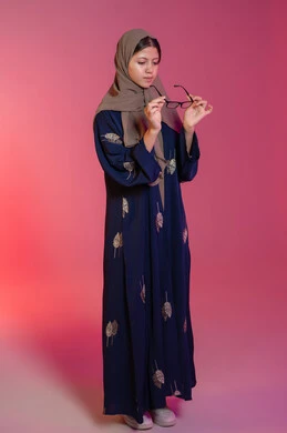 ارتداء النظارات الخاصة بضعف النظر, الاهتمام بالمظهر الخارجي، مفهوم ضعف البصر، بورتريه لامرأة عربية خليجية اماراتية محجبة ترتدي عباءة زرقاء تقوم باختيار نظارة ملائمة لملامح الوجه، تصحيح وتحسين الرؤية، متعة التسوق في الإمارات، خلفية وردية