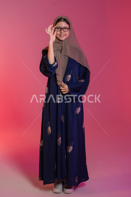 Correcting and improving vision, the pleasure of shopping in the Emirates, buying new eyeglasses, the concept of weak vision, a portrait of a smiling veiled Emirati Gulf Arab woman wearing a blue abaya choosing glasses that suit her facial features, pink background