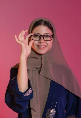 The concept of poor vision, buying new eyeglasses, portrait of a smiling veiled Emirati Gulf Arab woman wearing a blue abaya choosing glasses that suit her facial features, correcting and improving vision, the joy of shopping in the Emirates, pink background