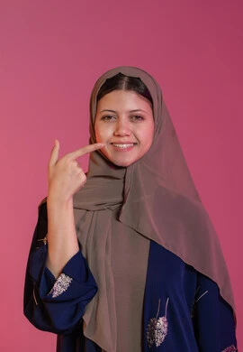 الإعتناء بنظافة وصحة الأسنان، الاهتمام بالمظهر الخارجي, بورتريه لامرأة عربية خليجية اماراتية محجبة مبتسمة ترتدي عباءة زرقاء تشير باصبع السبابة الى أسنانها, النظر الى الكاميرا بايماءات السعادة والسرور, تجنب التسوس وأمراض اللثة، خلفية وردية