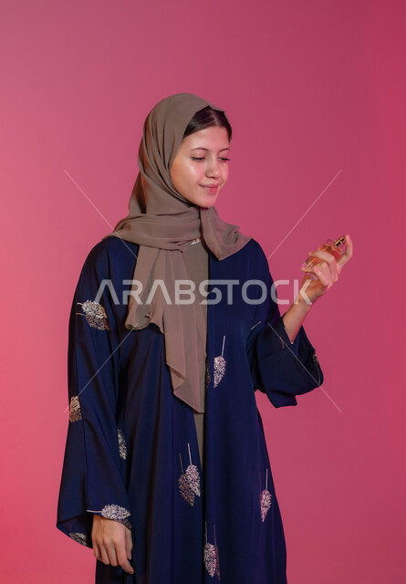 الشغف برش العطور, التجمل والتزيين النسائي, التسويق للمستحضرات النسائية, التطيب والتعطر بالعود والزيوت العطرية, مفهوم الأناقة والاهتمام بالمظهر الخارجي, بورتريه جانبي لامرأة عربية خليجية اماراتية محجبة مبتسمة ترتدي عباءة زرقاء تمسك زجاجة العطر، خلفية وردية