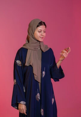 الشغف برش العطور, التجمل والتزيين النسائي, التسويق للمستحضرات النسائية, التطيب والتعطر بالعود والزيوت العطرية, مفهوم الأناقة والاهتمام بالمظهر الخارجي, بورتريه جانبي لامرأة عربية خليجية اماراتية محجبة مبتسمة ترتدي عباءة زرقاء تمسك زجاجة العطر، خلفية وردية