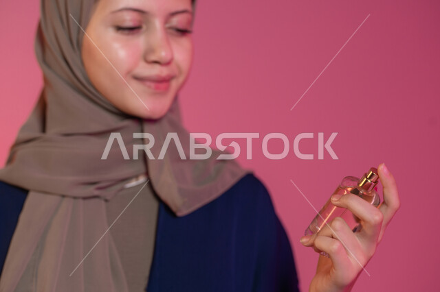 روائح عطرية متميزة، التسويق لمنتجات العطور والمستحضرات النسائية, التطيب والتعطر بالعود والزيوت العطرية, مفهوم الأناقة والاهتمام بالمظهر الخارجي, بورتريه جانبي لامرأة عربية خليجية اماراتية محجبة مبتسمة ترتدي عباءة زرقاء تقوم برش العطر، خلفية وردية