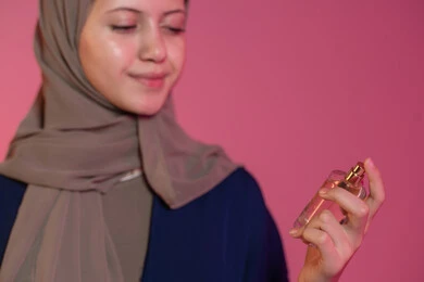 روائح عطرية متميزة، التسويق لمنتجات العطور والمستحضرات النسائية, التطيب والتعطر بالعود والزيوت العطرية, مفهوم الأناقة والاهتمام بالمظهر الخارجي, بورتريه جانبي لامرأة عربية خليجية اماراتية محجبة مبتسمة ترتدي عباءة زرقاء تقوم برش العطر، خلفية وردية
