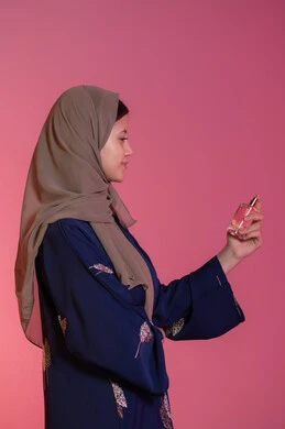 التجمل والتزيين النسائي, التسويق لمنتجات العطور والمستحضرات النسائية, التطيب والتعطر بالعود والزيوت العطرية, مفهوم الأناقة والاهتمام بالمظهر الخارجي, بورتريه جانبي لامرأة عربية خليجية اماراتية محجبة مبتسمة ترتدي عباءة زرقاء تقوم برش العطر، خلفية وردية