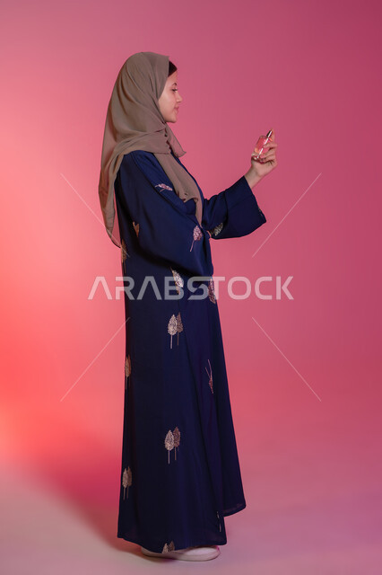 التسويق لمنتجات العطور والمستحضرات النسائية, التطيب والتعطر بالعود والزيوت العطرية, مفهوم الأناقة والاهتمام بالمظهر الخارجي, بورتريه جانبي لامرأة عربية خليجية اماراتية محجبة مبتسمة ترتدي عباءة زرقاء تقوم برش العطر، خلفية وردية