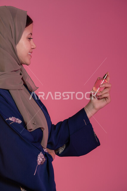 Perfumery and perfuming with oud and aromatic oils, marketing of perfumes and women's cosmetics, the concept of elegance and interest in outward appearance, a side portrait of a smiling veiled Emirati Gulf Arab woman wearing a blue abaya spraying perfume, pink background