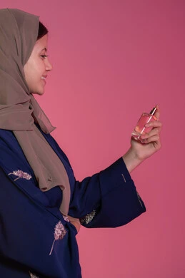 Perfumery and perfuming with oud and aromatic oils, marketing of perfumes and women's cosmetics, the concept of elegance and interest in outward appearance, a side portrait of a smiling veiled Emirati Gulf Arab woman wearing a blue abaya spraying perfume, pink background