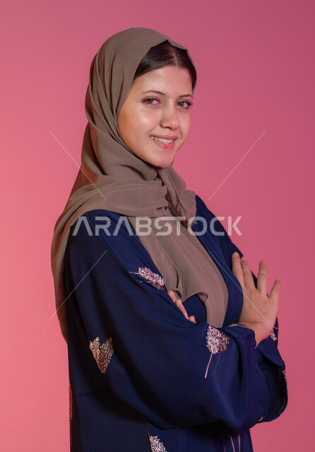 Expressions of self-confidence, interest in fashion and elegance, concept of taking care of one's appearance, looking at the camera with gestures of happiness and pleasure, side portrait of a smiling veiled Emirati Gulf Arab woman wearing a blue abaya, standing straight and balanced with her hands crossed, pink background