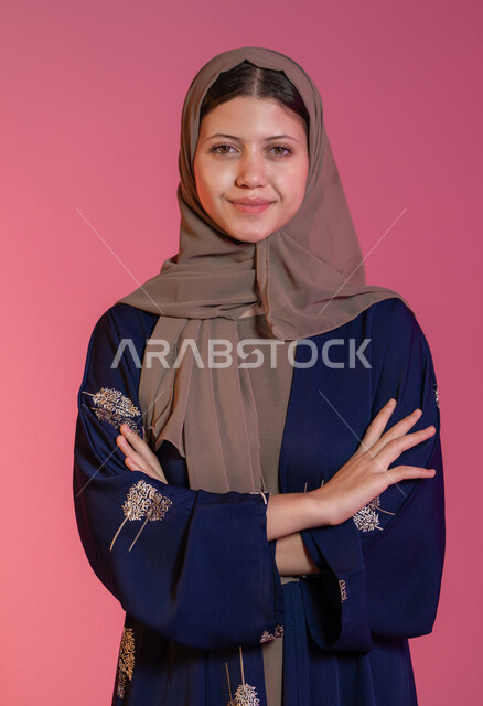 إيماءات الثقة بالنفس، الاهتمام بالمظهر الخارجي، مفهوم الانوثة والاناقة والنعومة،  النظر إلى الكاميرا بإيماءات السعادة والسرور, بورتريه مقرب  لامرأة عربية خليجية اماراتية محجبة مبتسمة ترتدي عباءة زرقاء تقف باستقامة واعتدال وأيدي متقاطعة, خلفية وردية