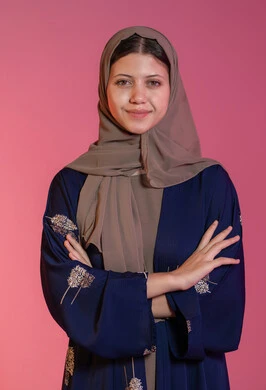 إيماءات الثقة بالنفس، الاهتمام بالمظهر الخارجي، مفهوم الانوثة والاناقة والنعومة،  النظر إلى الكاميرا بإيماءات السعادة والسرور, بورتريه مقرب  لامرأة عربية خليجية اماراتية محجبة مبتسمة ترتدي عباءة زرقاء تقف باستقامة واعتدال وأيدي متقاطعة, خلفية وردية