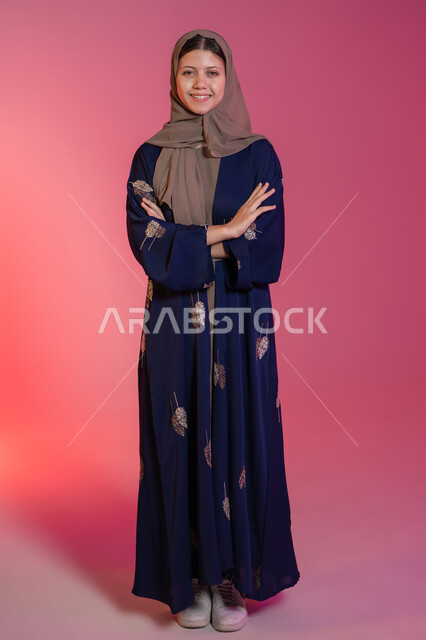 Concern for elegance and outward appearance, expressions of confidence and self-esteem, the concept of femininity and softness, looking at the camera with gestures of happiness and pleasure, a portrait of a smiling veiled Emirati Gulf Arab woman wearing a blue abaya, standing straight and balanced with her hands crossed, pink background