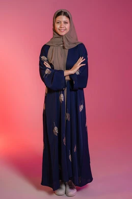 Concern for elegance and outward appearance, expressions of confidence and self-esteem, the concept of femininity and softness, looking at the camera with gestures of happiness and pleasure, a portrait of a smiling veiled Emirati Gulf Arab woman wearing a blue abaya, standing straight and balanced with her hands crossed, pink background