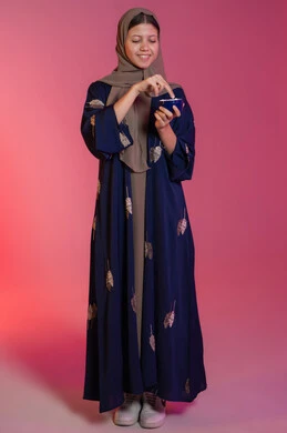 مفهوم العناية بنضارة وتحسين البشرة، استعمال مكون تجميلي، الاهتمام بالمظهر الخارجي للنساء، بورتريه مقرب لامرأة عربية خليجية اماراتية محجبة مبتسمة ترتدي عباءة زرقاء تحمل بيدها علبة كريم الترطيب، خلفية وردية