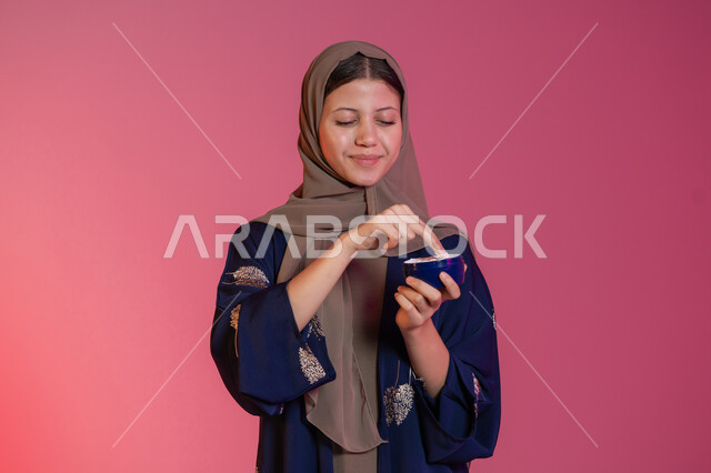 استعمال مكون تجميلي، مفهوم العناية بنضارة وتحسين البشرة، الاهتمام بالمظهر الخارجي للنساء، بورتريه مقرب لامرأة عربية خليجية اماراتية محجبة مبتسمة ترتدي عباءة زرقاء تحمل بيدها علبة كريم الترطيب، خلفية وردية