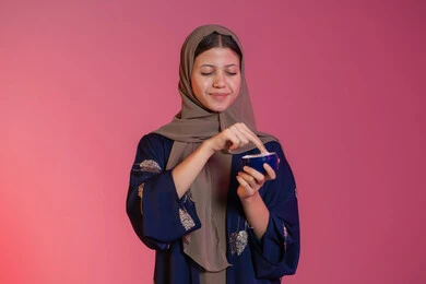 استعمال مكون تجميلي، مفهوم العناية بنضارة وتحسين البشرة، الاهتمام بالمظهر الخارجي للنساء، بورتريه مقرب لامرأة عربية خليجية اماراتية محجبة مبتسمة ترتدي عباءة زرقاء تحمل بيدها علبة كريم الترطيب، خلفية وردية