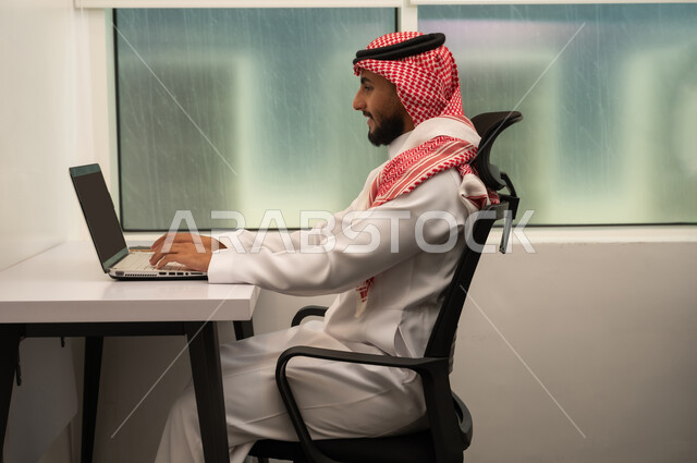 The concept of remote business management, the use of modern devices and advanced technologies, office professions and jobs, an Arab Gulf Saudi man wearing traditional attire and a headscarf in front of a laptop, using the computer to send emails.