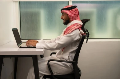 The concept of remote business management, the use of modern devices and advanced technologies, office professions and jobs, an Arab Gulf Saudi man wearing traditional attire and a headscarf in front of a laptop, using the computer to send emails.