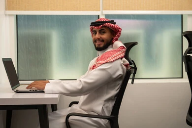 Planning and managing the company, the concept of achieving tasks, using modern devices and advanced technologies, office professions and jobs, an Arab Gulf Saudi man wearing traditional attire and a headscarf, using a laptop, looking at the camera with gestures of happiness and joy.