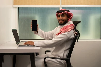 Performing job tasks through mobile, using a computer to send emails, integrating modern and advanced technologies with work, administrative office professions and jobs, a Saudi Gulf Arab man wearing traditional attire and a headscarf using a laptop and mobile phone, looking at the camera with expressions of happiness.