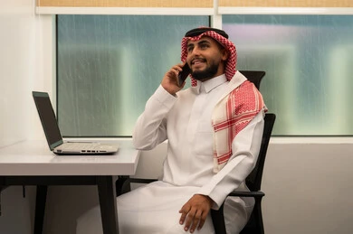 الاتصال عبر الجوال لإجراء مكالمة،  دراسه المشروع ومراجعه التقرير السنوي، رجل عربي خليجي سعودي يرتدي الثوب التقليدي و الشماغ و يقوم باستخدام اللابتوب و الموبايل, دمج التقنيات الحديثة و المتطورة مع العمل