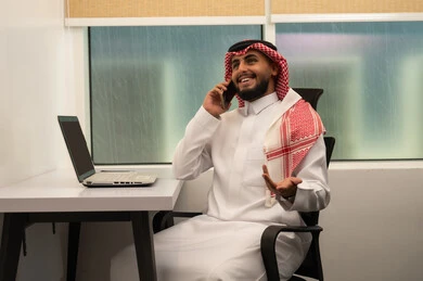 إجراء مكالمة صوتية عبر الهاتف المحمول، دراسه المشروع ومراجعه التقرير السنوي، رجل عربي خليجي سعودي يرتدي الثوب التقليدي و الشماغ و يقوم باستخدام اللابتوب و الموبايل,  دمج التقنيات الحديثة و المتطورة مع العمل