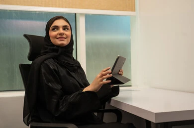A professional meeting was held online, a comfortable work environment in Saudi companies, women's office professions and jobs, a smiling Arab Gulf Saudi woman wearing a black abaya holding a tablet and looking away, the concept of achieving work.
