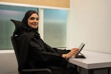 انجاز و ادارة المهام باستخدام جهاز لوحي, تنظيم اجتماع إلكتروني، بيئة عمل مريحة في الشركات السعودية, مهن و وظائف مكتبية نسائية, إمرأة عربية خليجية سعودية مبتسمة ترتدي عباءة سوداء و تستخدم التابلت, ايماءات السعادة و السرور