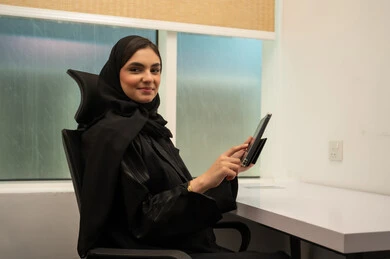 ايماءات السعادة و السرور، تنظيم اجتماع عبر الإنترنت، بيئة عمل مريحة في الشركات السعودية, مهن و وظائف مكتبية نسائية, إمرأة عربية خليجية سعودية مبتسمة ترتدي عباءة سوداء و تمسك جهاز التابلت, مفهوم انجاز الاعمال