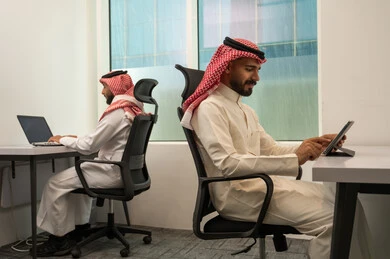 Meeting planning and task scheduling, collaboration in completing work, strategic plans to achieve goals, lifestyle in Saudi companies, two Arab Gulf Saudi men wearing traditional thob and ghutrah sitting in the office and using advanced devices.