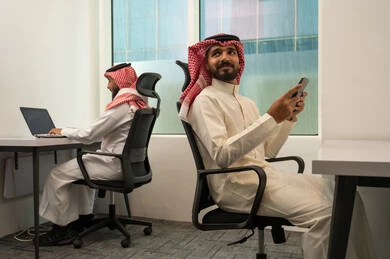 بيئة تعاونية مكتبية, التشارك في إنجاز الاعمال, الخطط الاستراتيجية لتحقيق الأهداف، نمط الحياة في الشركات السعودية، رجلان عربيان خليجيان سعوديان يرتديان الثوب التقليدي و الشماغ يجلسون في المكتب و يقومان باستخدام الأجهزة المتطورة