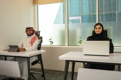 The spirit of collaboration and public conversations, strategic plans to achieve goals, the lifestyle in Saudi companies, a Gulf Arab Saudi team consisting of a man wearing traditional attire and a shemagh and a woman in a hijab wearing a black abaya, accomplishing tasks through a laptop and a tablet.