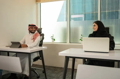 Gestures of happiness and integration, the spirit of collaboration and public conversations, strategic plans to achieve goals, the lifestyle in Saudi companies, an Arab Gulf Saudi team consisting of a man wearing traditional attire and a headscarf and a woman in a black abaya, accomplishing tasks through a laptop and tablet.
