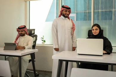 نقاشات عمل و محادثات عامة، الخطط الاستراتيجية لتحقيق الأهداف،  نمط الحياة في الشركات السعودية، فريق عربي خليجي سعودي مكون من رجلين يرتديان الثوب التقليدي و الشماغ و سيدة محجبة ترتدي العباءة السوداء, انجاز المهام من خلال اللابتوب و التابلت, تعزيز الروح التشاركية