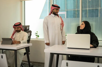 Training employees on work policy, supervising projects in the office, promoting a collaborative spirit, distributing tasks required to develop the company's goals, lifestyle in Saudi companies, a Gulf Arab Saudi team consisting of two men wearing traditional thobes and ghutras and a veiled woman wearing a black abaya, completing tasks using laptops and tablets.
