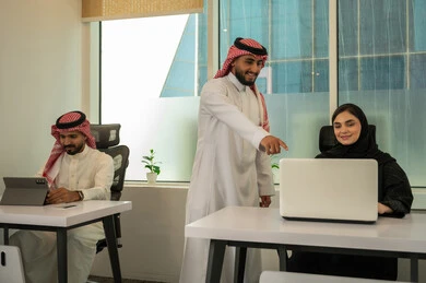 نمط الحياة في الشركات السعودية، تدريب الموظفين على سياسات و مهام العمل لتحقيق الأهداف في المكتب داخل مقر الشركة،  فريق عربي خليجي سعودي مكون من رجلين يرتديان الثوب التقليدي و الشماغ و سيدة محجبة ترتدي العباءة السوداء, انجاز المهام من خلال اللابتوب و التابلت, الاشارة الى الكمبيوتر