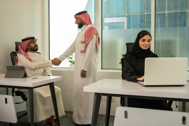 مصافحة الزملاء داخل المكتب, التعاون و متابعة الاعمال, نمط الحياة في الشركات السعودية، تدريب الموظفين على سياسات العمل لتحقيق الأهداف داخل مقر الشركة، فريق عربي خليجي سعودي مكون من رجلين يرتديان الثوب التقليدي و الشماغ و سيدة محجبة ترتدي العباءة السوداء, انجاز المهام من خلال اللابتوب و التابلت
