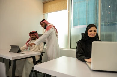 Supervising projects at the workplace, enhancing the spirit of collaboration, distributing the tasks required to develop the company's goals, the lifestyle in Saudi companies, an Arab Gulf Saudi team consisting of two men wearing traditional thob and shemagh and a veiled woman wearing a black abaya, completing tasks using a laptop and tablet, referring to the tablet.
