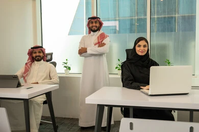 Collaboration and business follow-up, the lifestyle in Saudi companies, training employees on work policies to achieve goals in the office within the company headquarters, a Gulf Arab Saudi team consisting of two men wearing traditional thob and ghutrah and a veiled woman wearing a black abaya, completing tasks using a laptop and tablet, standing upright with arms crossed.