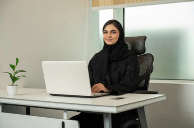 إتمام المهام عبر الإنترنت، إدارة شؤون الأعمال و تنظيمها عبر الكمبيوتر، استخدام جهاز تقني حديث و متطور،  سيدة عربية خليجية سعودية محجبة ترتدي عباءة سوداء و أمامها جهاز اللابتوب, النظر الى الكاميرا بايماءات السعادة و السرور