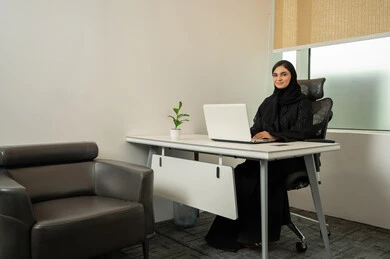 مهن و وظائف مكتبية نسائية, إمرأة عربية خليجية سعودية مبتسمة ترتدي عباءة سوداء و أمامها جهاز اللابتوب, النظر الى الكاميرا بايماءات السعادة و السرور, إتمام المهام عبر الإنترنت، إدارة شؤون الأعمال و تنظيمها عبر الكمبيوتر، استخدام جهاز تقني حديث و متطور